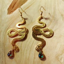 Load image into Gallery viewer, Gold-plated coiled snake drop earrings with textured scale detail and a green crystal charm, laid on a wooden surface - Let's Jam jewellery collection