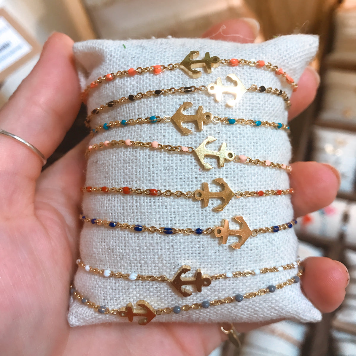 A row of gold-tone chain bracelets with anchor charms and colourful beaded accents, pinned to a cushion and held up to show the full collection of colours - Let's Jam jewellery collection