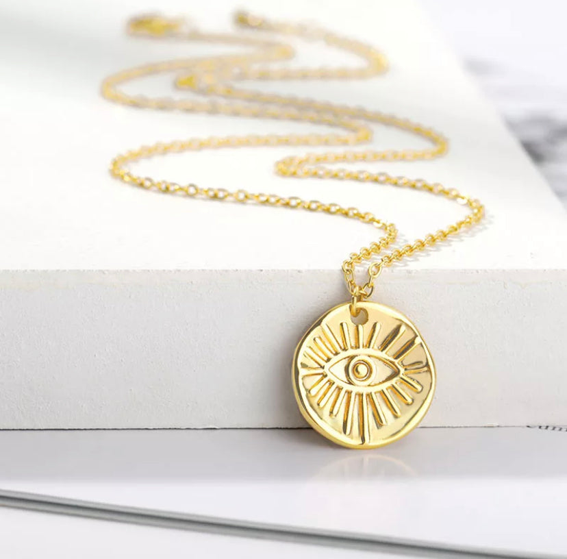 Gold-plated round pendant necklace engraved with a protective evil eye design, laid on a stack of books beside a delicate chain - Let's Jam jewellery collection