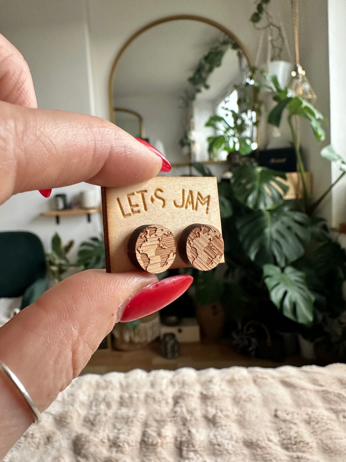 Hand holding a Let's Jam branded card with a pair of round wooden stud earrings engraved with a world map design, plants in the background - Let's Jam jewellery collection