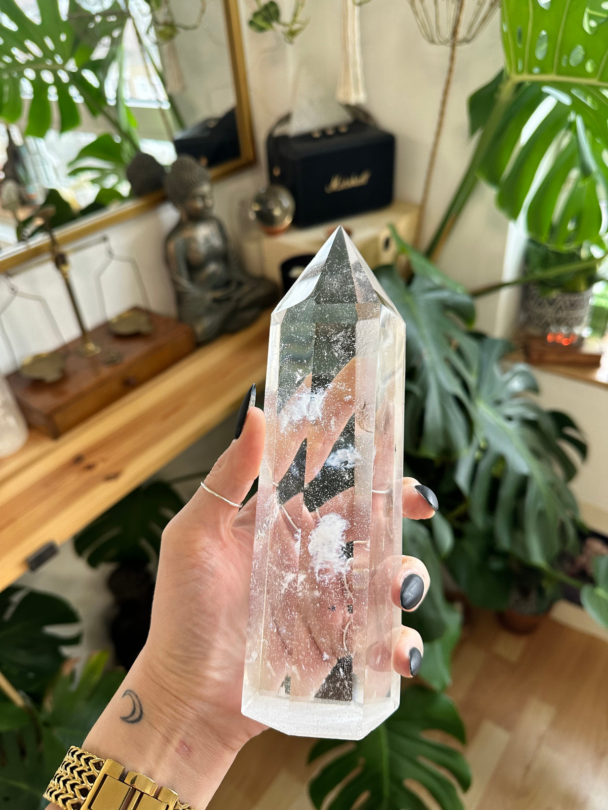 Hand holding a clear quartz crystal tower point with dramatic internal fracture patterns, houseplants and a small statue in the background - Let's Jam crystal collection