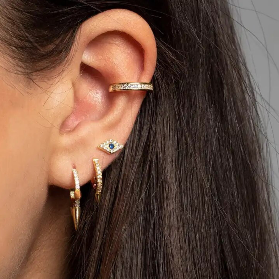 Close-up of a woman's ear styled with a textured gold-plated ear cuff, a small blue evil eye stud earring, and layered gold hoop earrings with crystal detail - Let's Jam jewellery collection