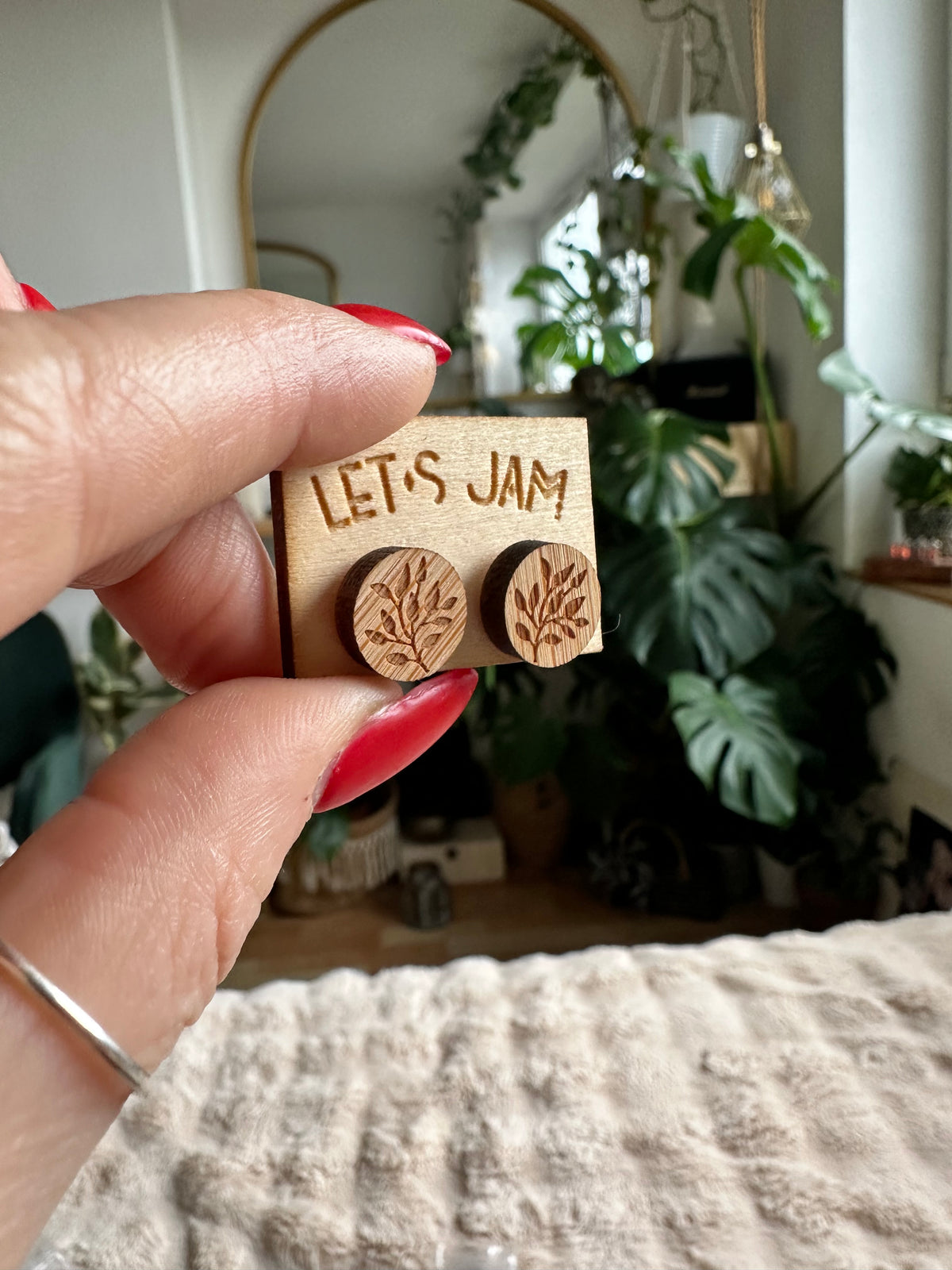 Hand holding a Let's Jam branded card with a pair of round wooden stud earrings engraved with a large leaf design, plants in the background - Let's Jam jewellery collection