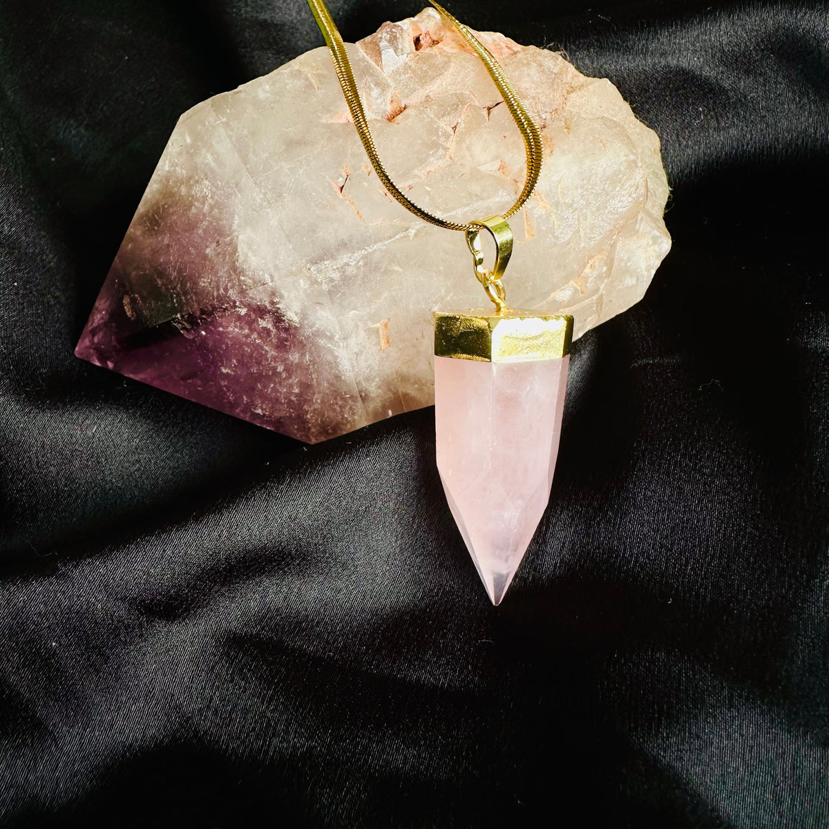 Rose quartz crystal point pendant on a gold snake chain necklace, displayed against a natural rose quartz crystal on black fabric - Let's Jam jewellery collection