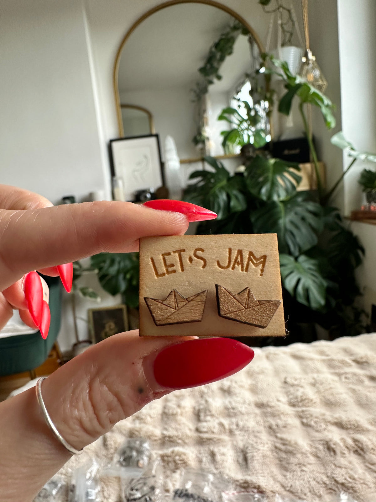 Hand holding a Let's Jam branded card with a pair of wooden stud earrings engraved with a paper-boat design, plants in the background - Let's Jam jewellery collection