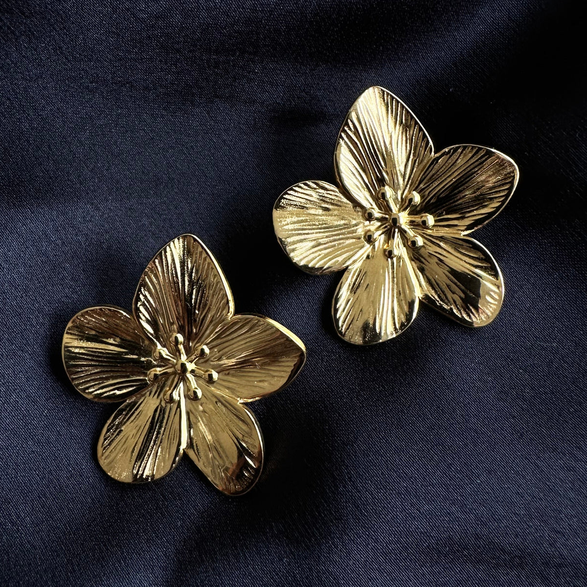 Pair of gold-plated stainless steel flora flower stud earrings with textured petal detailing, laid on dark navy fabric - Let's Jam jewellery collection