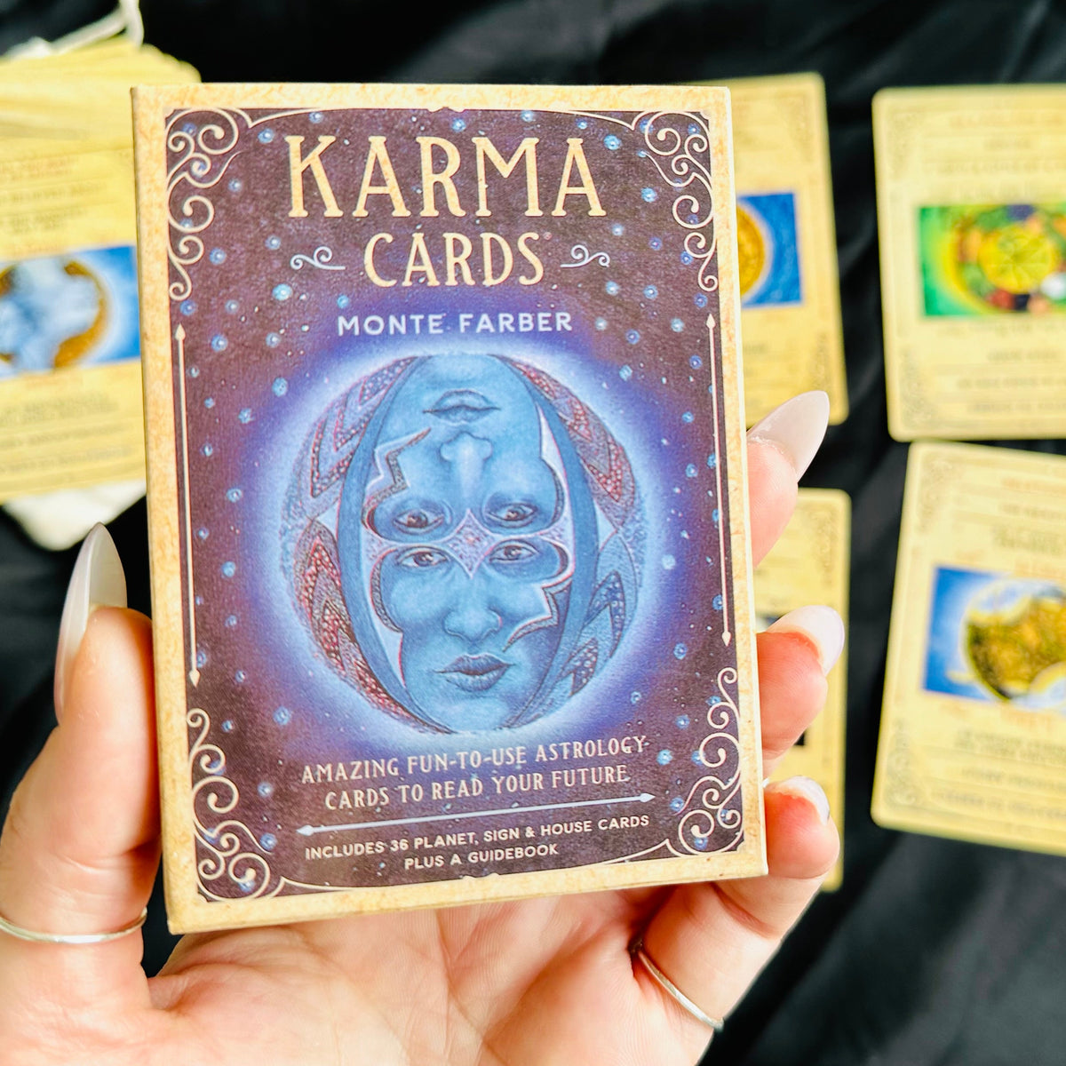 Hand holding the Karma Cards astrology oracle deck box with illustrated cosmic face design, fanned out beside colourful zodiac and planet cards on black fabric - Let's Jam collection