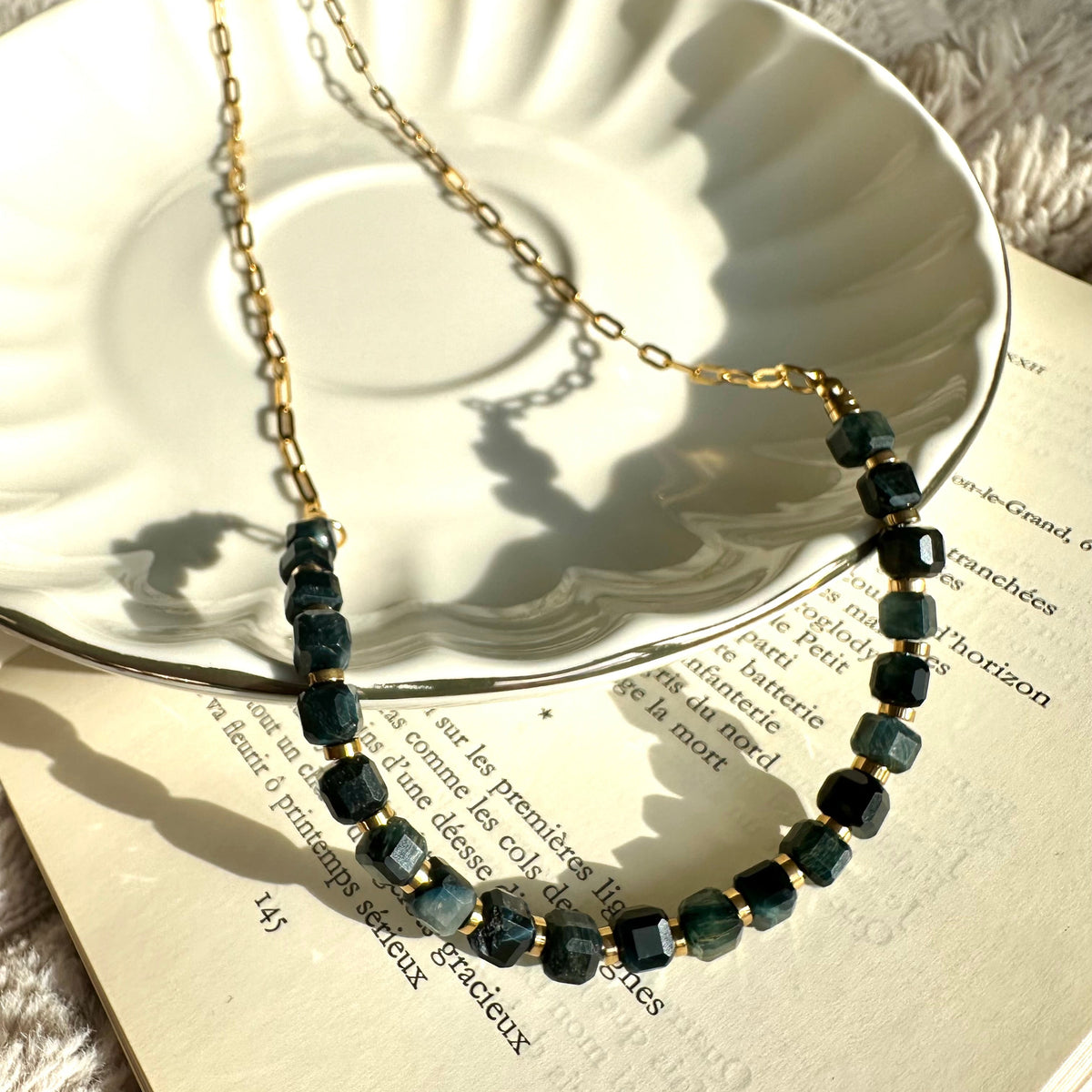 Black tiger's eye beaded necklace with 18k gold-plated stainless steel chain, laid on an open book beside a white plate - Let's Jam jewellery collection