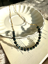 Load image into Gallery viewer, Black tiger's eye beaded necklace with 18k gold-plated stainless steel chain, laid on an open book beside a white plate - Let's Jam jewellery collection
