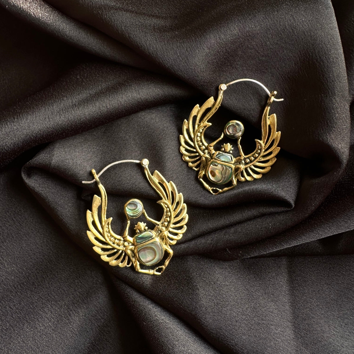 Pair of handcrafted brass hoop earrings shaped like winged scarab beetles with blue abalone shell inlay, displayed on dark satin fabric - Let's Jam jewellery collection