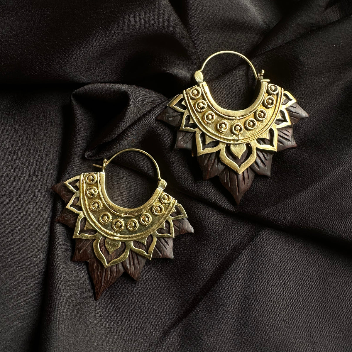 Pair of ornate gold-tone hoop earrings with an intricately carved dark wood lotus flower petal fringe, displayed on dark satin fabric - Let's Jam jewellery collection