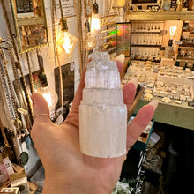 Load image into Gallery viewer, A hand holding a tiered white crystal tower in a cluttered, vintage-style jewelry shop with warm hanging light bulbs and display counters in the background.
