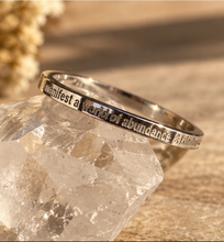 Load image into Gallery viewer, A silver band ring engraved with the quote 'I Manifest a world of abundance &amp; kindness' resting on a clear quartz crystal against a warm, sandy background.
