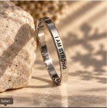 Load image into Gallery viewer, A polished silver-toned band ring engraved with the words 'I AM STRONG' in black lettering, leaning against a textured beige stone on a sandy surface with warm sunlight and leaf shadows.

