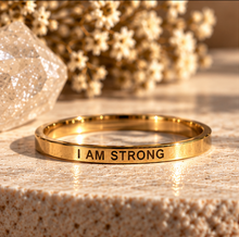 Load image into Gallery viewer, A polished gold ring engraved with the words 'I AM STRONG' in black lettering, resting on a textured beige stone surface with dried flowers and a crystal in the background.
