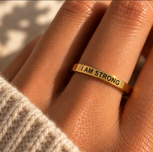 Load image into Gallery viewer, A gold band ring engraved with the words 'I AM STRONG' in black lettering, worn on a person's finger. The shot is taken in warm, direct sunlight with a beige knit sweater visible at the bottom left.
