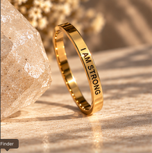 Load image into Gallery viewer, A gold ring engraved with the words 'I AM STRONG' in black lettering, standing upright on a textured surface next to a clear crystal.
