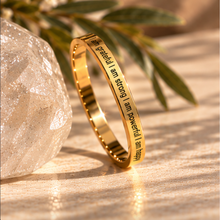 Load image into Gallery viewer, A polished gold ring engraved with the affirmations 'I am grateful, I am strong, I am powerful, I am happy' in black lettering, styled on a textured beige surface with a crystal and green leaves in the background.
