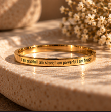 Load image into Gallery viewer, A polished gold ring with engraved positive affirmations sits on a textured beige ceramic dish with dried white flowers in the background.
