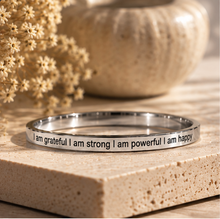 Load image into Gallery viewer, A polished silver metal ring with black engraved text reading 'I am grateful I am strong I am powerful I am happy', displayed on a textured beige stone block with dried flowers and a blurred ceramic bowl in the background.
