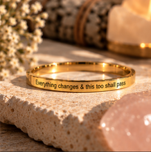 Load image into Gallery viewer, A gold ring engraved with the quote 'Everything changes &amp; this too shall pass' in black lettering, resting on a textured stone slab with warm, boho-style background props.
