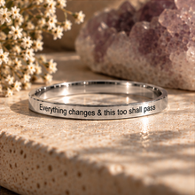 Load image into Gallery viewer, A polished silver ring engraved with the quote 'Everything changes &amp; this too shall pass' in black lettering. The ring is displayed on a textured beige stone surface, styled with dried white flowers and a purple crystal cluster in the background.
