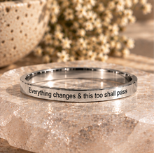 Load image into Gallery viewer, A polished silver metal ring engraved with the quote 'Everything changes &amp; this too shall pass', displayed on a textured stone slab with dried flowers in the background.
