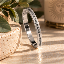Load image into Gallery viewer, A polished silver-toned metal ring with engraved black text reading 'Everything changes &amp; this too shall pass', displayed standing upright on a textured beige surface with decorative leaves and a sun-themed candle in the background.

