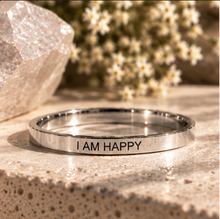 Load image into Gallery viewer, A polished silver-toned metal ring engraved with the words 'I AM HAPPY' in black lettering, resting on a textured beige stone surface with a blurred background of small white flowers and a crystal.
