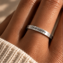 Load image into Gallery viewer, A silver band ring engraved with the words 'I AM HAPPY' worn on a finger, shown in a close-up lifestyle shot with a cream knit sleeve visible.
