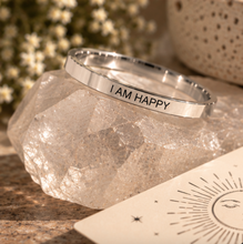 Load image into Gallery viewer, A polished silver ring engraved with the words 'I AM HAPPY' in black capital letters, resting on a faceted clear crystal. The scene is styled with a warm, bohemian background featuring dried flowers, a textured ceramic pot, and a cream-colored card with a sun illustration.
