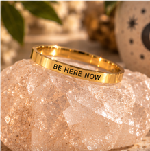 Load image into Gallery viewer, A polished gold ring engraved with the words 'BE HERE NOW' in black lettering, displayed on a faceted clear crystal cluster with a warm, boho-style background.

