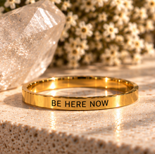 Load image into Gallery viewer, A polished gold ring engraved with the words 'BE HERE NOW' in black capital letters, resting on a textured beige stone surface with a blurred crystal and small white flowers in the background.
