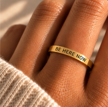 Load image into Gallery viewer, A gold band ring engraved with the words 'BE HERE NOW' in black lettering, worn on a person's finger. The shot is a close-up with warm, natural lighting and a soft-focus background.
