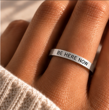 Load image into Gallery viewer, A close-up of a hand wearing a silver band ring engraved with the words 'BE HERE NOW' in black capital letters, with a cream knit sleeve visible at the bottom left.
