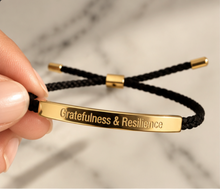 Load image into Gallery viewer, A close-up of a hand holding a black braided cord bracelet with a polished gold ID bar engraved with the words 'Gratefulness &amp; Resilience'. The background is a soft-focus marble surface.
