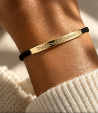 Load image into Gallery viewer, A close-up of a person's wrist wearing a black cord bracelet with a polished gold metal bar engraved with the words 'Gratitude &amp; Resilience'. The person is wearing a cream-colored knit sweater, and the background is softly blurred with warm sunlight.
