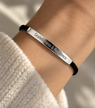 Load image into Gallery viewer, A close-up of a person's wrist wearing a silver bar bracelet engraved with 'Gratitude &amp; Resilience' on a black cord bracelet, styled with a cream knit sleeve.
