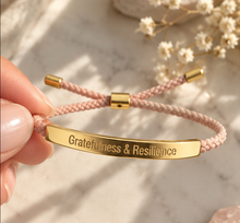 Load image into Gallery viewer, A close-up of a hand holding a pink braided cord bracelet with a polished gold-tone ID bar engraved with the words 'Gratitude &amp; Resilience'. The background is a soft-focus marble surface with dried white flowers.
