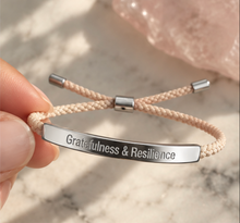 Load image into Gallery viewer, A close-up of a hand holding a beige braided cord bracelet with a polished silver bar engraved with the words 'Gratefulness &amp; Resilience'. The background is a soft-focus marble surface with a pink crystal.
