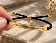 Load image into Gallery viewer, A close-up of a hand holding a black braided cord bracelet with a polished gold bar engraved with the quote, "I am exactly where I am supposed to be". The bracelet has gold-toned metal end caps and sliding adjustment beads.
