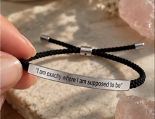 Load image into Gallery viewer, A close-up of a hand holding a black braided cord bracelet with a silver metal ID plate engraved with the quote: 'I am exactly where I am supposed to be'. The background features a textured light stone and pink crystal clusters.

