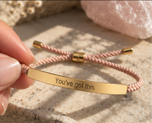 Load image into Gallery viewer, A close-up of a hand holding a pink braided cord bracelet with a gold-tone ID bar engraved with the words 'You've got this'. The background consists of a textured stone surface with a pink crystal.
