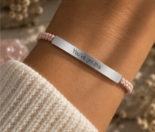 Load image into Gallery viewer, A close-up of a person's wrist wearing a silver bar bracelet engraved with 'You've got this' in black lettering, paired with a light pink braided cord bracelet, styled with a cream knit sleeve and a blurred pink stone background.
