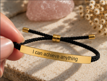 Load image into Gallery viewer, A close-up of a hand holding a black braided cord bracelet with a polished gold-tone rectangular plate engraved with the phrase 'I can achieve anything'. The bracelet features gold-tone sliding beads and end caps.

