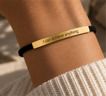 Load image into Gallery viewer, A close-up of a person's wrist wearing a black cord bracelet with a polished gold rectangular bar engraved with the motivational phrase 'I can achieve anything'.

