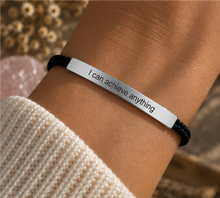 Load image into Gallery viewer, A close-up of a person's wrist wearing a silver bar bracelet engraved with the phrase 'I can achieve anything' in black lettering. The bracelet is secured with a black braided cord and is styled with a cream-colored knit sweater, with a soft-focus background of dried flowers and a pink crystal.
