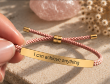 Load image into Gallery viewer, A close-up of a hand holding a pink braided cord bracelet with a gold bar engraved with the phrase 'I can achieve anything'. The background features a textured beige stone and a pink crystal.
