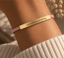 Load image into Gallery viewer, A close-up of a person's wrist wearing a pink braided cord bracelet with a rectangular gold metal nameplate engraved with the phrase 'I can achieve anything'.
