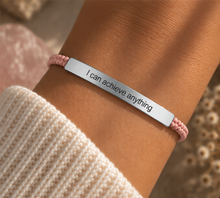 Load image into Gallery viewer, A close-up of a person's wrist wearing a silver bar bracelet engraved with the phrase 'I can achieve anything'. The bracelet is flanked by a light pink braided cord, and the person is wearing a cream-colored knit sweater.

